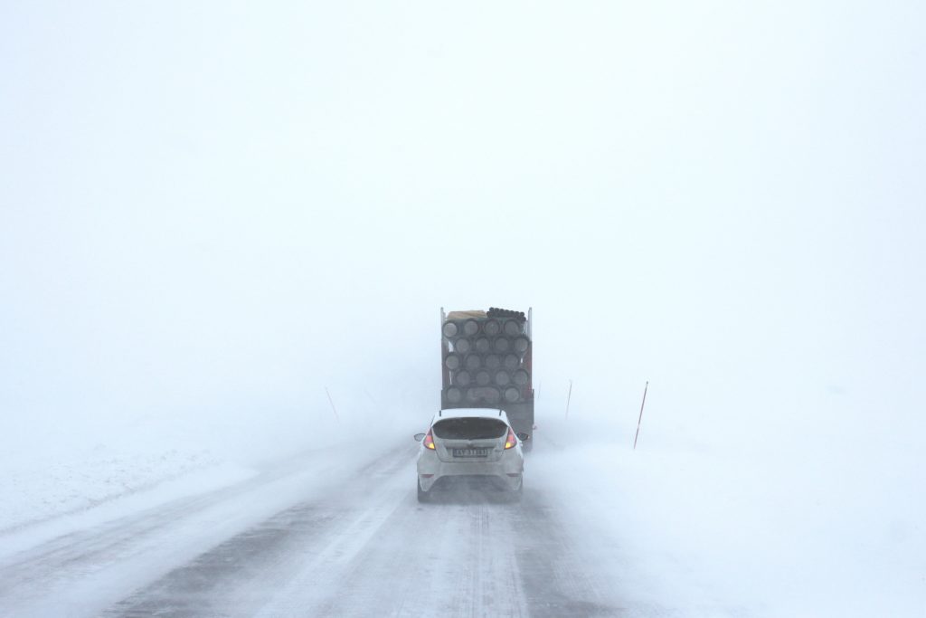 log truck and car following closely behind in a snowstorm
