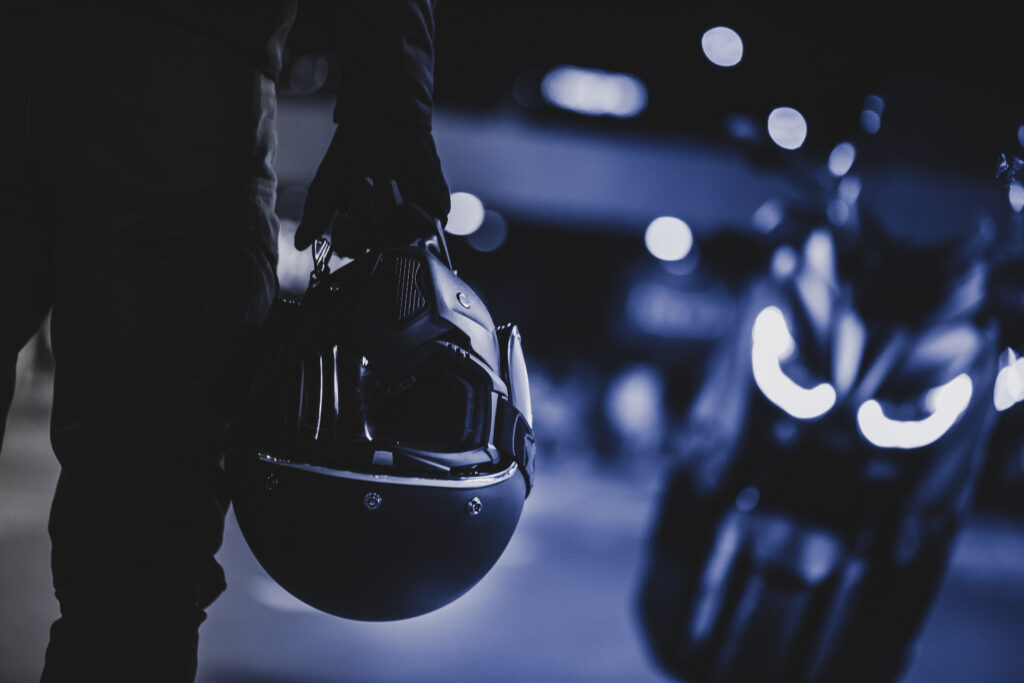A motorcyclist with a protective helmet in his hand next to his motorcycle. Taken at night. Photograph