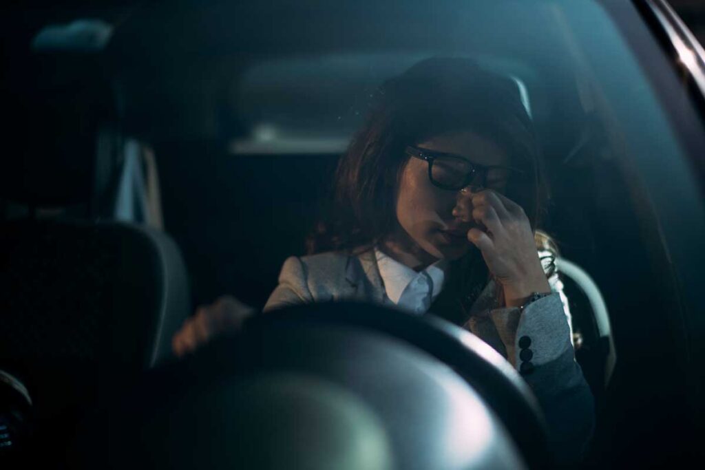 woman driving in her car, point of view from the driver window, holding her head under her glasses signaling a headache after fall back savings
