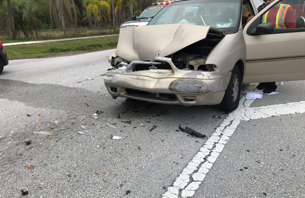 A tan car with a smashed front end from a car accident sits in the middle of the road surrounded by broken glass and debris from the accident in Clearwater, FL.