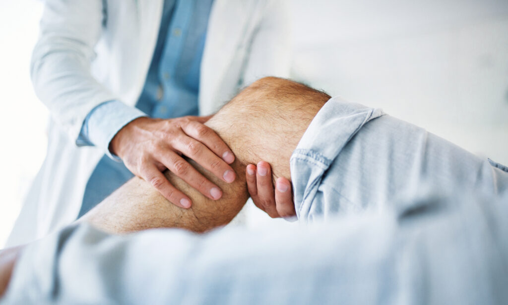 Closeup side view of early 30's unrecognizable doctor examining a knee of a senior gentleman during an appointment. The doctor is gently touching the tendons around the knee and the knee cap and evaluating after an injury