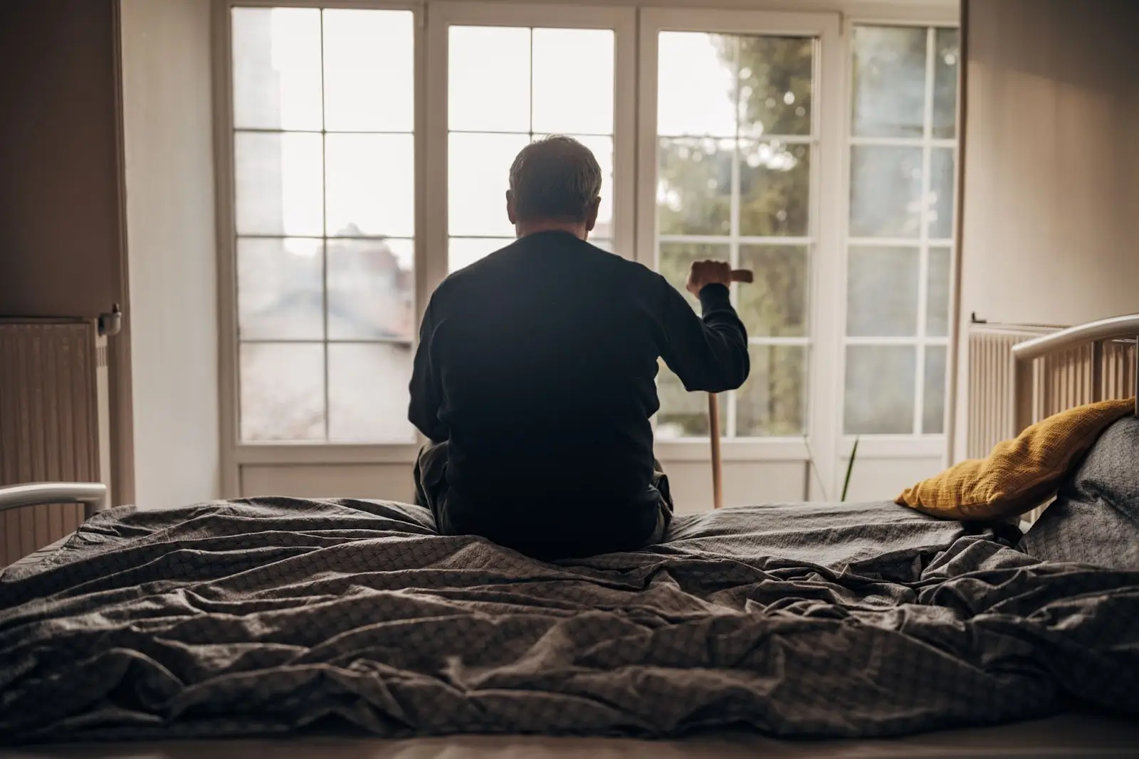 An older man sits on the edge of his bed, holding his cane and looking out the window.