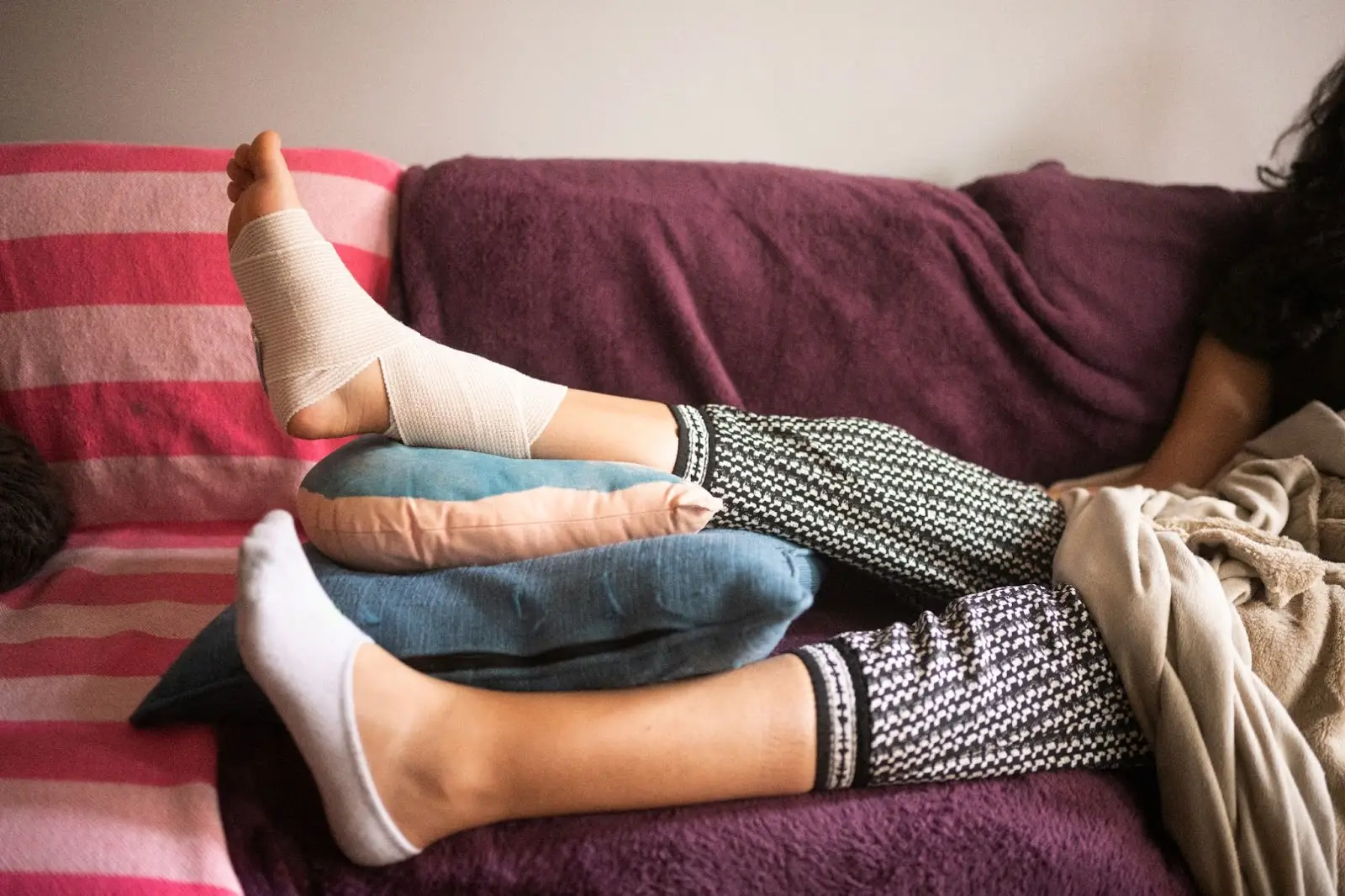 a victim of an accident lies on their couch. with their bandaged ankle elevated on pillows.