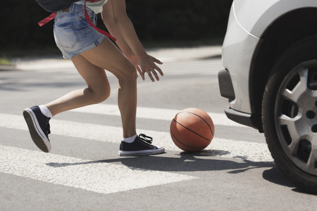 a young person reaches for a basketball in a crosswalk as a white car approaches from their right.
