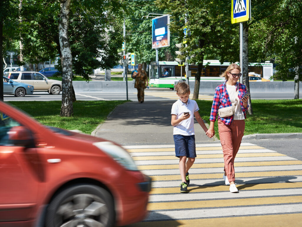 A mother and child walking through a crosswalk in Florida both looking at their phones, coming close to being hit by a red car driving through.