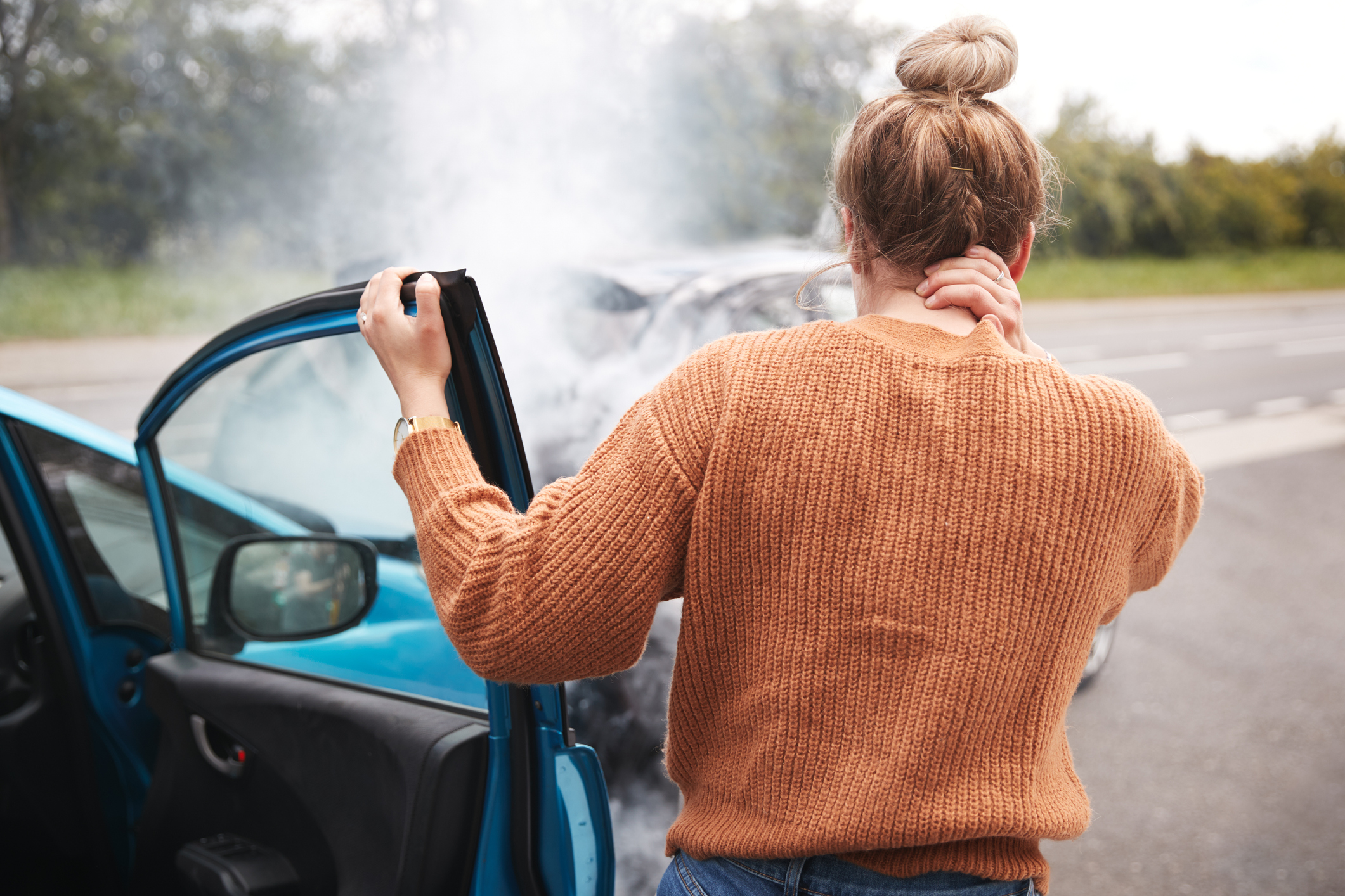 A passenger steps out of a vehicle after an accident holding her neck while she looks at the damage to the cars.