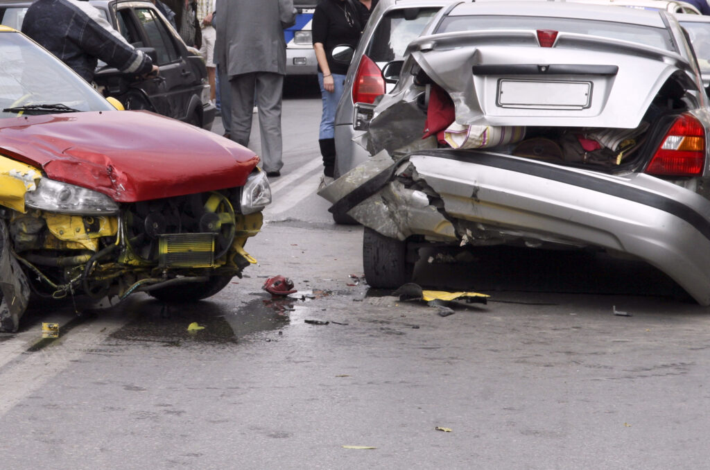 Two cars are damaged after a car accident. Debris is scattered on the ground, and bystanders are looking on.