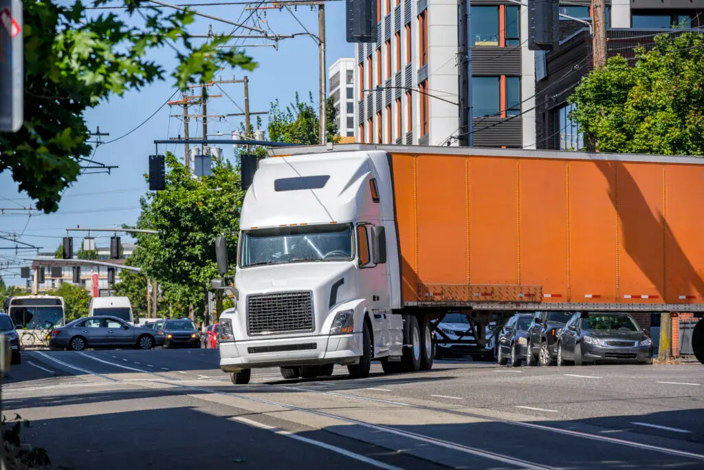 An orange semi-truck takes a wide turn on a busy urban street.