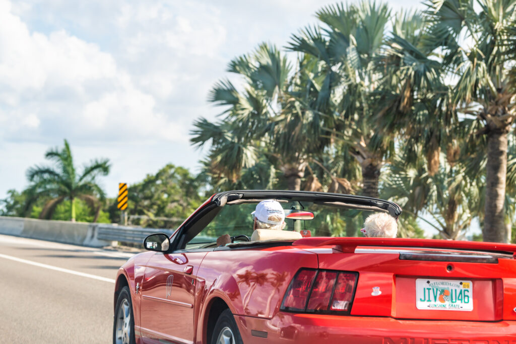 An older couple drives down a Florida highway in a convertible red sports car.