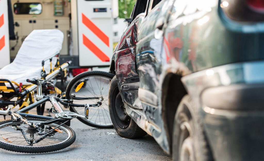 A car with damage to its side panel after an accident with a cyclist. An ambulance and a stretcher are in the background.