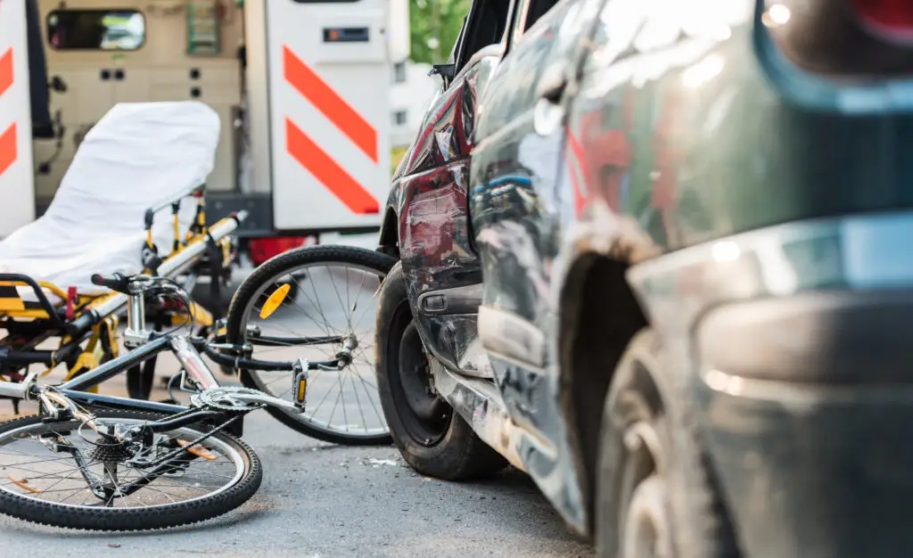 A car with damage to its side panel after an accident with a cyclist. An ambulance and a stretcher are in the background. 