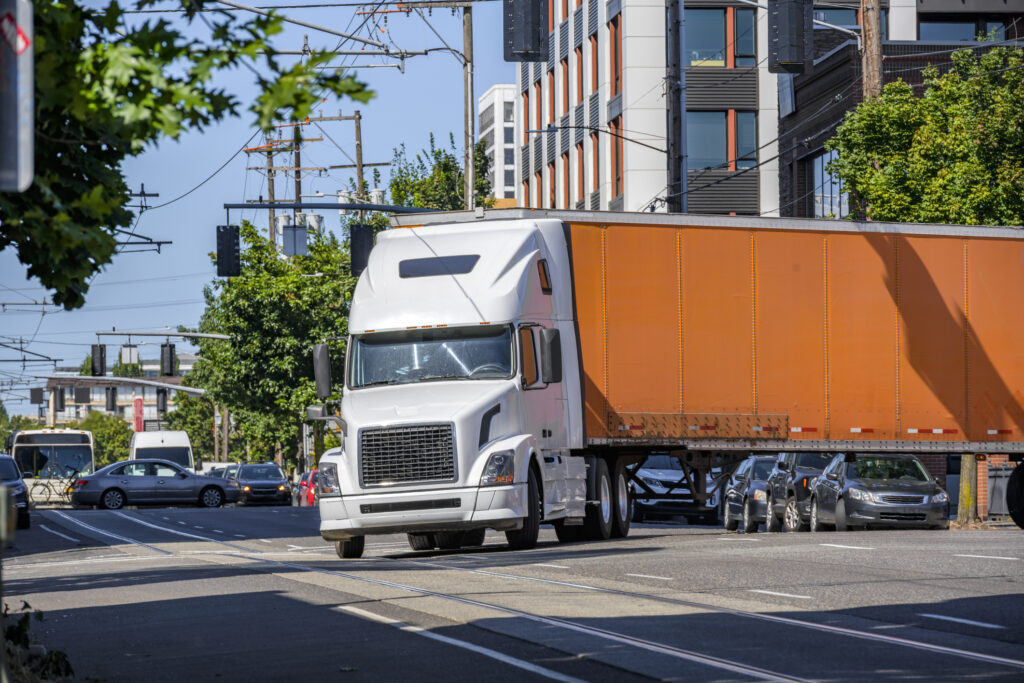 A large semi truck with a large orange trailer makes a wide turn into traffic at an intersection.