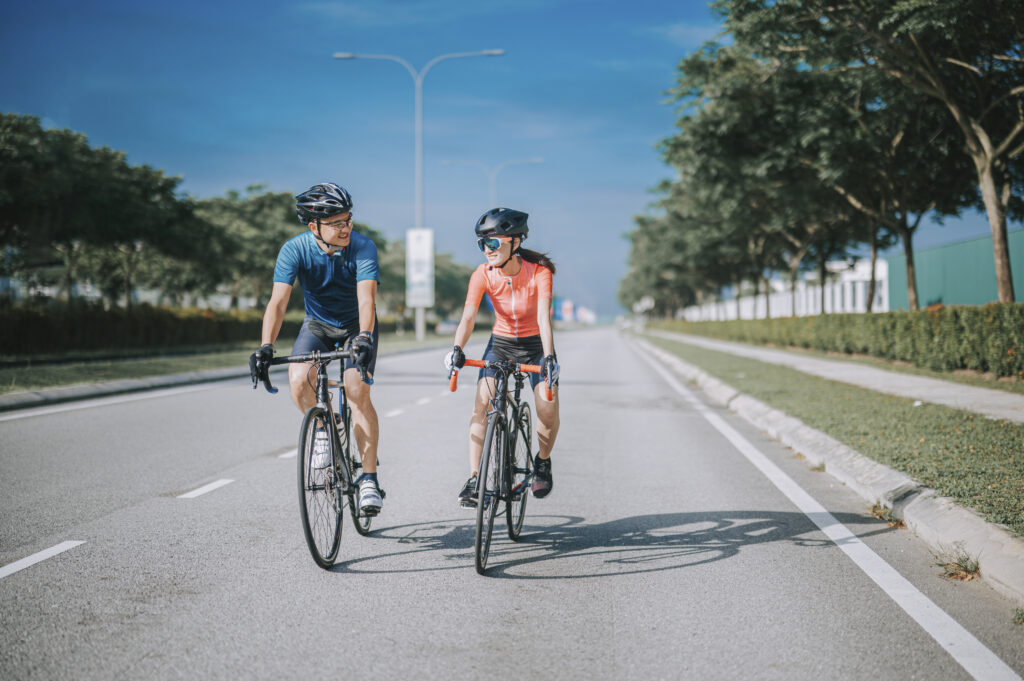 A man and a woman wearing helmets riding their bicycles on a tree-lined road.