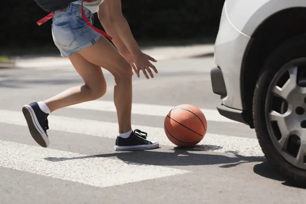 A young girl chases a basketball onto the road in a crosswalk in front of an oncoming car.