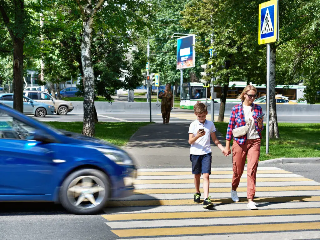 A mother and son cross the street holding hands and looking at their smartphones while a blue automobile approaches them on the road, potentially leading to a pedestrian accident with severe injuries.